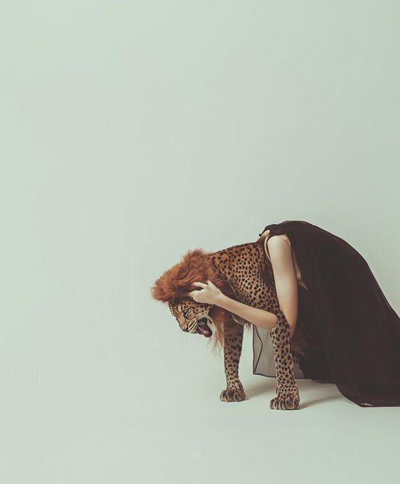 An artistic photograph of a person in a dark dress bending forward while embracing a cheetah against a pale mint-green studio background.