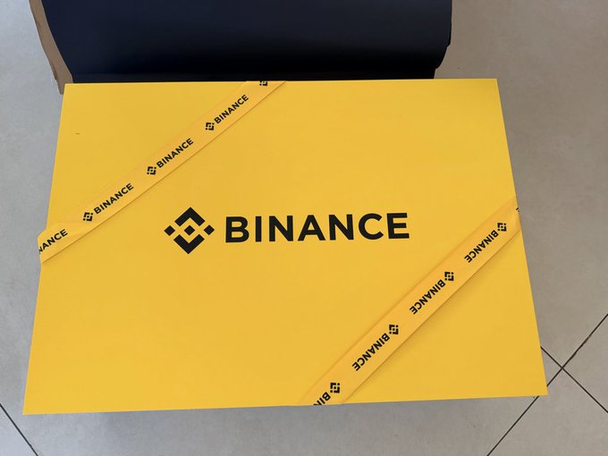 A bright yellow Binance-branded envelope or package with the Binance logo, placed on a tiled floor.