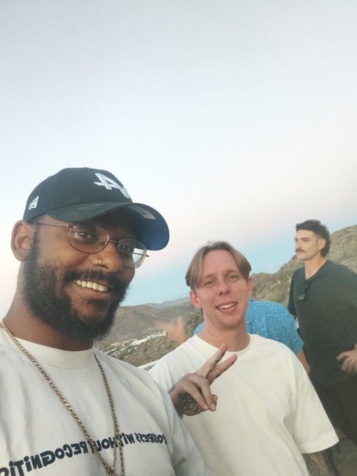 A selfie of two smiling men outdoors with hills in the background; the man in front wears a black 'LA' Dodgers cap and glasses, while his companion in a white shirt makes a peace sign.