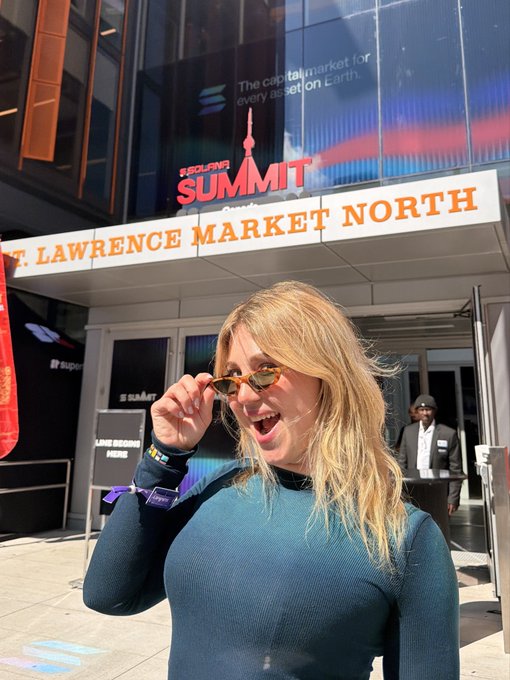 A blonde woman in a teal long-sleeve top and sunglasses poses outside the 'St. Lawrence Market North' building, with a 'TORONTO SUMMIT' banner visible above the entrance.