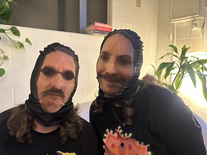 The image features two people wearing masks that resemble animal heads or human faces with googly eyes. They appear to be posing together, possibly as part of a costume event or a fun gathering. The masks are made from fabric and have a unique design, adding an element of humor and creativity to the scene.