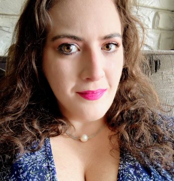 A selfie of a woman with long curly brown hair, wearing pink lipstick and a delicate pearl necklace, looking directly at the camera.