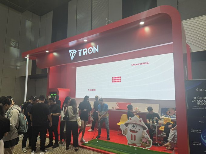 A trade show or convention booth for 'TRON' featuring a large red structure with the Tron logo, an LED screen displaying graphics, and various attendees interacting in the space.