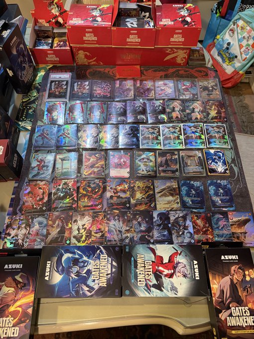 An expansive collection of Azuki TCG "Gates Awakened" trading cards is displayed on a playmat, with unopened booster boxes and starter decks featuring Azuki and other characters arranged in the foreground, celebrating the set's release day.