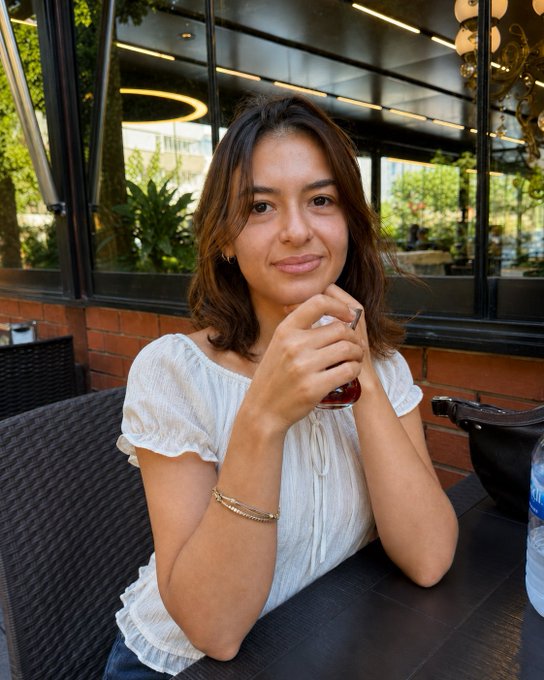 A young woman with shoulder-length brown hair wearing a white blouse, sitting at an outdoor café table and smiling at the camera while holding a small glass.