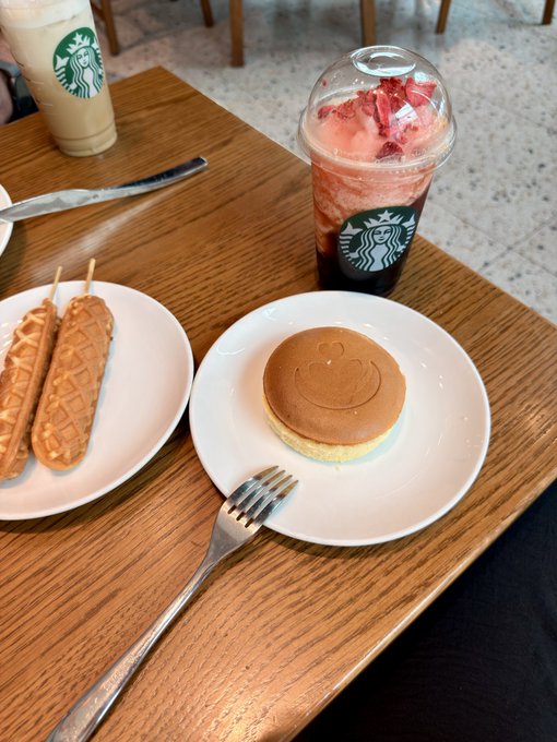 A Starbucks order on a wooden table featuring a latte, a strawberry drink, hot dog-shaped waffles, and a smiley face pancake with a fork.