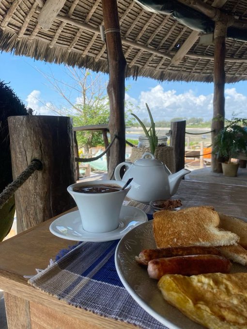 A photograph of a hearty breakfast on a wooden table at an open-air beachside hut, featuring coffee in a white cup, a teapot, toast, sausages, and an omelet with the ocean visible in the background.