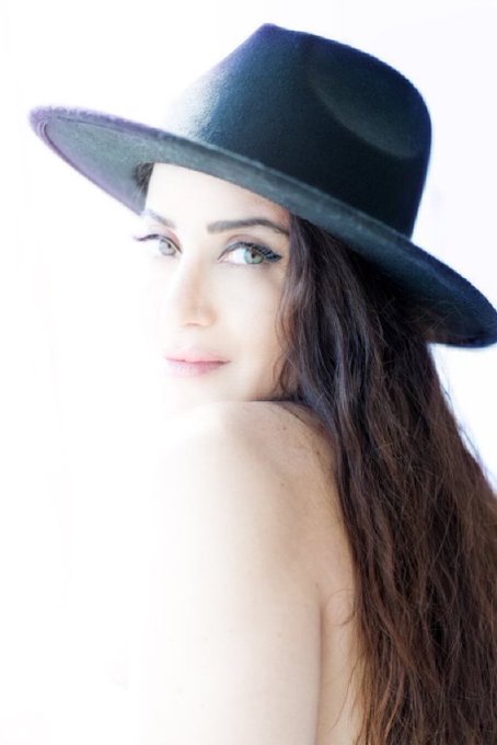 Portrait photograph of a woman with long dark hair wearing a black wide-brimmed hat, looking over her shoulder at the camera against a bright white background.