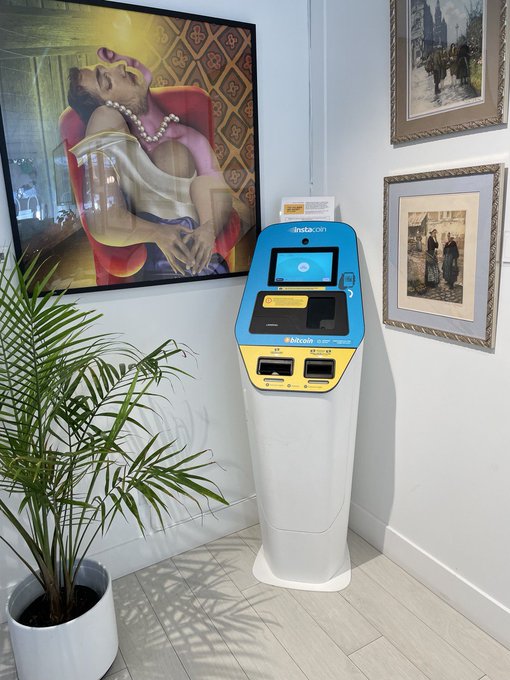A blue and yellow InstaKort Bitcoin ATM stands on a white tiled floor next to a potted palm plant, with framed artwork hanging on the wall behind it.