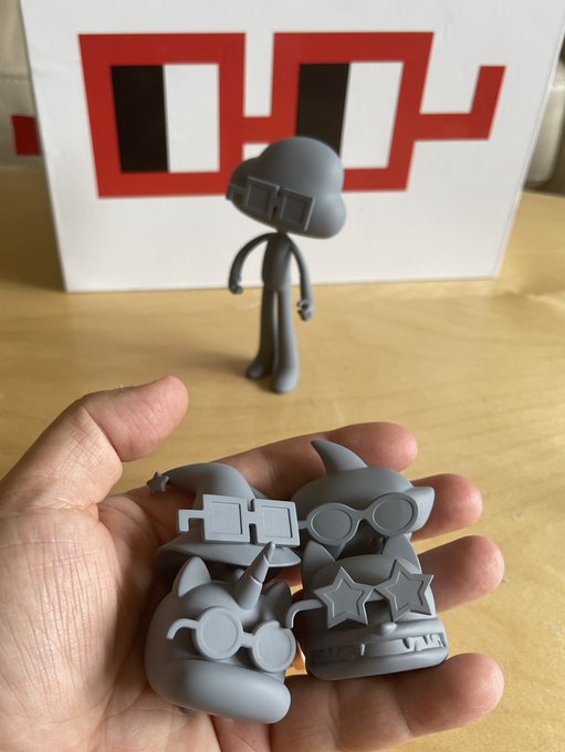 A hand holds four small gray 3D-printed figurines in front of a larger matching figurine, with a red and black "IP" logo box in the background.
