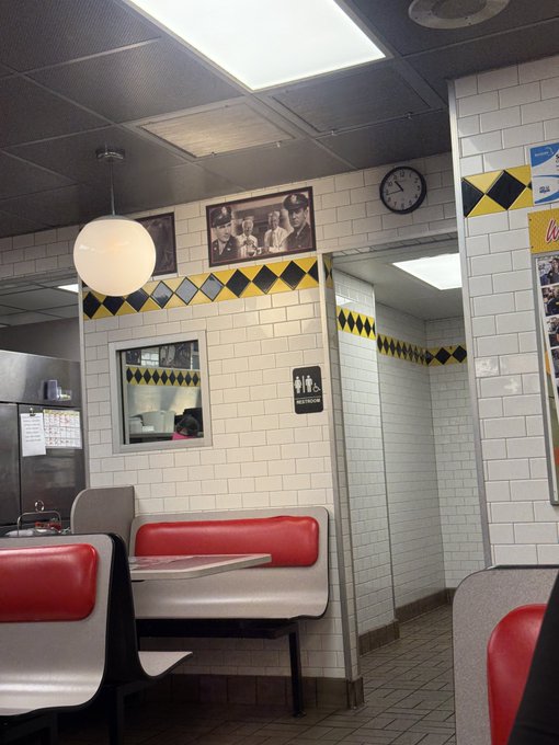 An interior shot of a retro-style diner featuring white subway tile walls with yellow and black checkered accents, red vinyl booths, a vintage wall clock, and a black-and-white framed photograph on the wall.