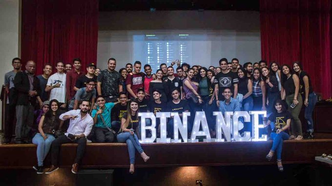 A large group of crypto enthusiasts gathers on a stage in Maracaibo for what appears to be a Binance community event, posing behind illuminated "BINANCE" letters while a data screen displays above them.
