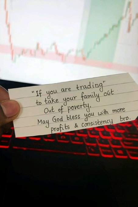 A hand holding a handwritten note that reads 'If you are trading to take your family out of poverty, May God bless you with more profits & consistency bro.' in front of a keyboard and monitor displaying a candlestick chart.