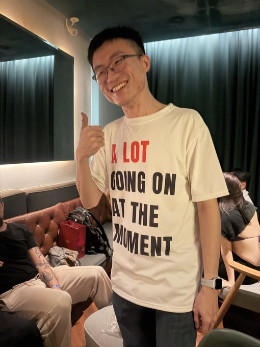 A cheerful young man wearing glasses and a t-shirt reading "A LOT GOING ON AT THE MOMENT" gives a thumbs-up, perfectly capturing the caption's nod to a well-known NFT community figure known for keeping tabs on the space.