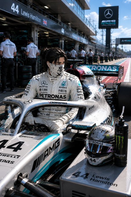 An anime-style illustration of a female driver in Petronas Mercedes racing gear sitting in car #44, with 'Lewis Hamilton' and a UK flag visible on the pit garage signage.