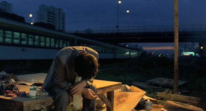 A man in a suit sits hunched over on a wooden table at dusk, appearing dejected, with a train passing on an overpass behind him.