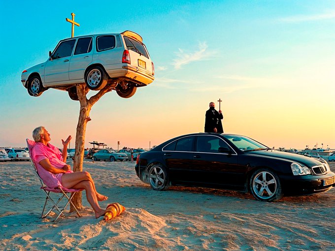 A surreal desert scene features a woman relaxing in a pink beach chair beneath a white SUV mounted on a wooden pole with a cross, while a man in black holds another cross near a black sedan.