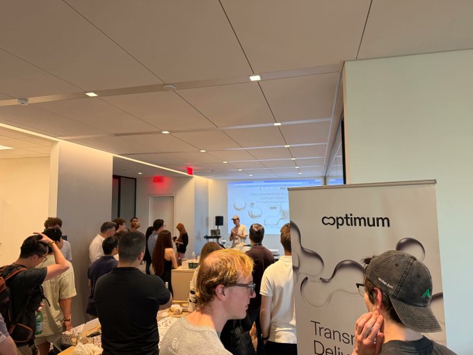 The image shows a group of people gathered around a table, with some standing behind a sign that says "Optimium". There are at least 12 people visible in the scene, including both men and women. They appear to be engaged in conversation or observing something on the table. The room has white walls and ceiling, creating an open and spacious atmosphere for the gathering.