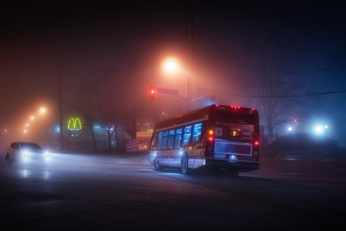 A foggy nighttime city street scene featuring a city bus with route number 7, illuminated by a streetlamp and traffic light, with a McDonald's golden arches sign visible in the background.