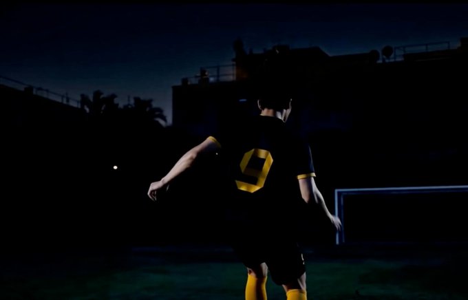 A young person seen from behind stands on a soccer field at night, wearing a black jersey with a yellow number 9.