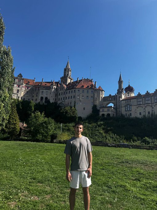 A young man in a gray t-shirt and white shorts poses on a grassy field with a large historic castle complex on a hill behind him.