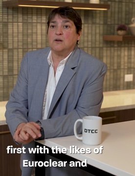 The image shows a man wearing a gray suit sitting at a table with a coffee cup, possibly a DTCC Euroclear mug. The background features a tiled wall that appears to be part of a kitchen or dining area. There are no other people in the scene, and the focus is on the man's attire and the coffee cup he holds.