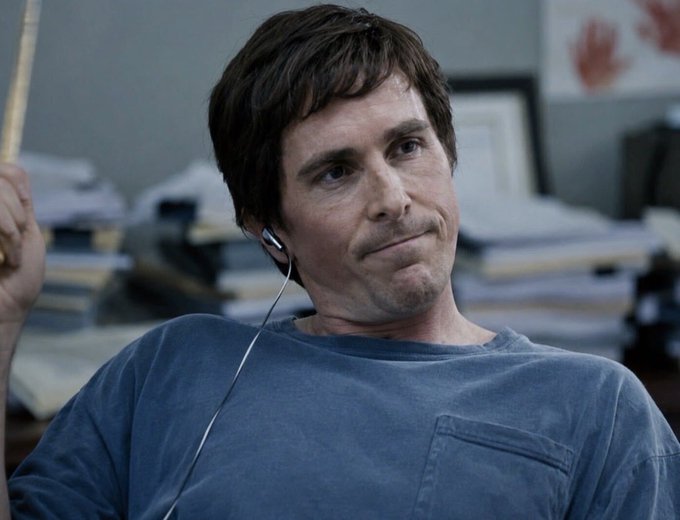 A man with brown hair, wearing earbuds and a grey shirt, sits in an office with stacks of papers behind him.