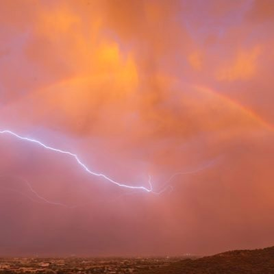 A dramatic lightning bolt streaking across an orange and pink sunset sky over a darkened landscape.