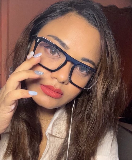 A selfie of a young woman wearing black-framed glasses, red lipstick, and blue manicured nails, holding her hand up to her face.