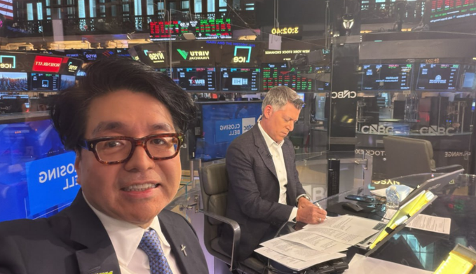 A selfie of a man in a suit and glasses at the CNBC trading floor, with another man working at a desk in the background.