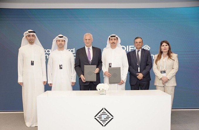 A group of six people, including men in traditional Arab white robes and others in business attire, stand behind a white podium displaying a logo, with two men holding what appear to be certificates or documents.