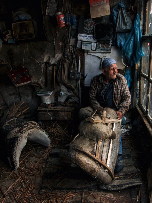 An elderly man wearing a blue cap and plaid shirt sits in a cluttered, rustic workshop surrounded by tools and materials, with a large animal pelt or hide draped over an object in front of him.