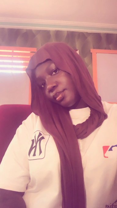 A selfie of a person wearing a maroon hijab and a white t-shirt with logos, posing in a room with orange and patterned walls.