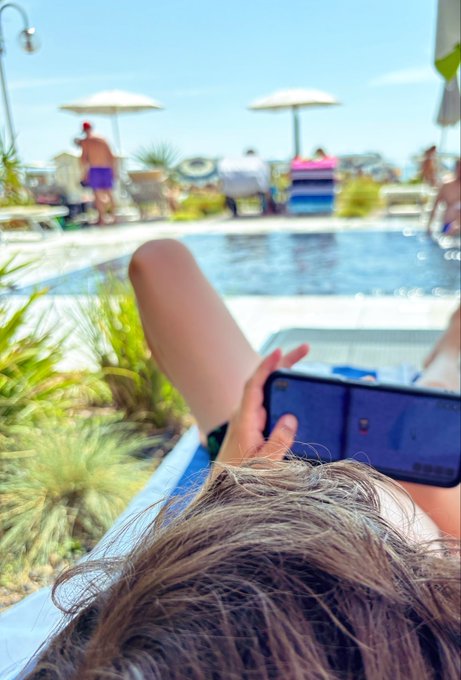A first-person view from a poolside lounge chair shows the top of a person's head and a hand holding a smartphone, with a swimming pool, umbrellas, and other guests in the sunny background.