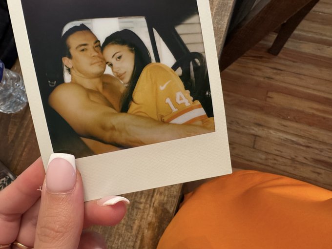 A hand holds a Polaroid photo of a couple posing together, with the woman wearing a yellow jersey bearing the number "14." The caption suggests a playful attempt to cover part of her face with a lollipop or candy, adding humor to the shared memory.