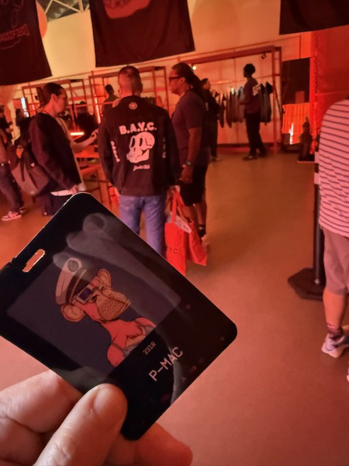 A hand holds up a black card featuring a cartoon character with goggles and the text 'P-MAC 219', with people browsing in a clothing store in the background, including one wearing a jacket with 'B.A.Y.C' and a skull logo.