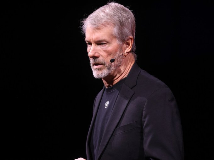 A man with gray hair and a beard, wearing a black suit jacket, speaking into a headset microphone against a dark background.