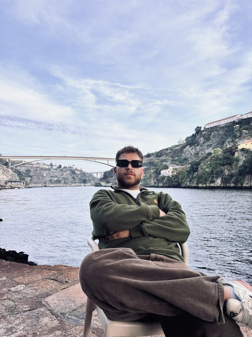 A man in sunglasses and a green hoodie sits cross-legged in a plastic chair by the water, with a bridge and hillside buildings visible in the background, projecting a relaxed, confident "good morning" vibe that matches the post's request to drop $SOL addresses.
