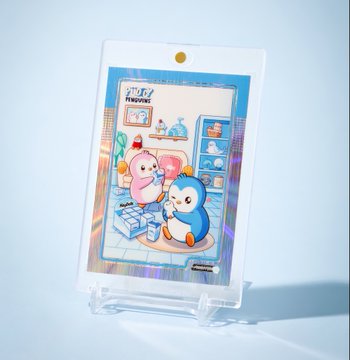 A holographic card or photo card featuring two cute penguin-like characters (Pi-chan and Pengin) in a bathroom scene, still in its plastic protective sleeve on a display stand.