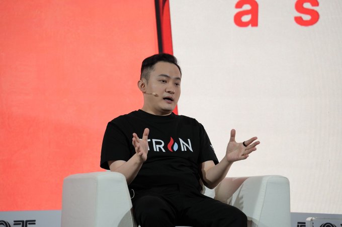Justin Sun, wearing a black TRON t-shirt, speaks at a DAS conference, gesturing with both hands while seated on a white chair against a red and white backdrop. His comments highlight that while agentic payments could align AI through incentives, current adoption remains limited due to insufficient AI capability.