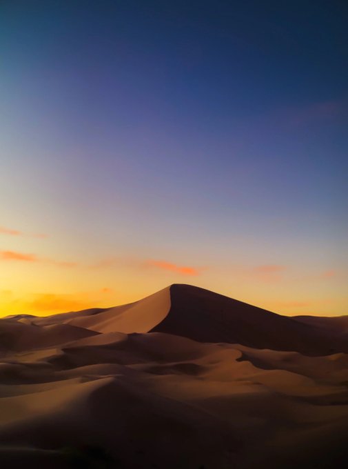 A desert landscape featuring rolling sand dunes silhouetted against a vibrant sunset sky with warm orange and yellow hues transitioning to blue above.