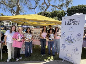 A group of young women and a woman pose together at an outdoor event booth with a yellow canopy, standing next to a promotional banner reading 'YOUR ALL CAMPUS CONCIERGE' from Visible Magic, with informational pamphlets displayed on the table.