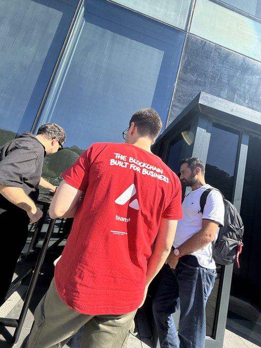 Three young men stand near a glass building, with one in the foreground wearing a red t-shirt reading 'THE BLOCKCHAIN BUILT FOR BUSINESS' with a logo and 'team?' text.