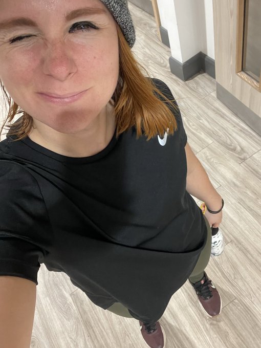 A woman winks while taking a gym selfie, wearing a black Nike t-shirt, gray beanie, and athletic sneakers, with "Gy-M" likely referencing her gym session.