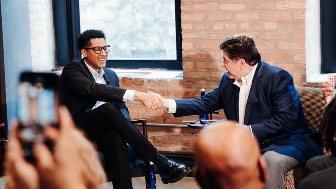 Two men in suits shaking hands and smiling during what appears to be a public event or interview, with an audience in the foreground.