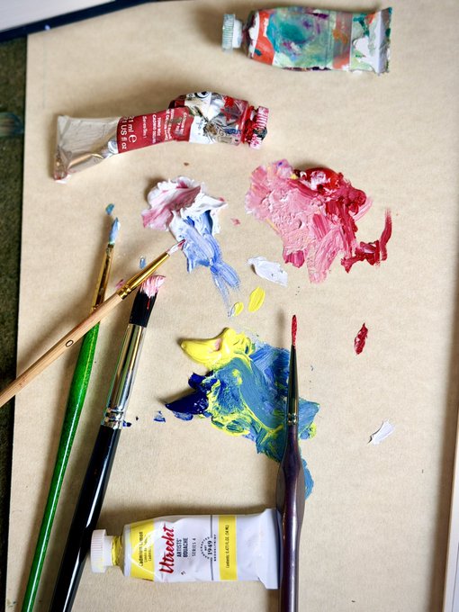 An overhead view of an artist's workspace featuring a palette covered in smears of red, blue, and yellow paint, along with paintbrushes and tubes of oil paint on brown paper.