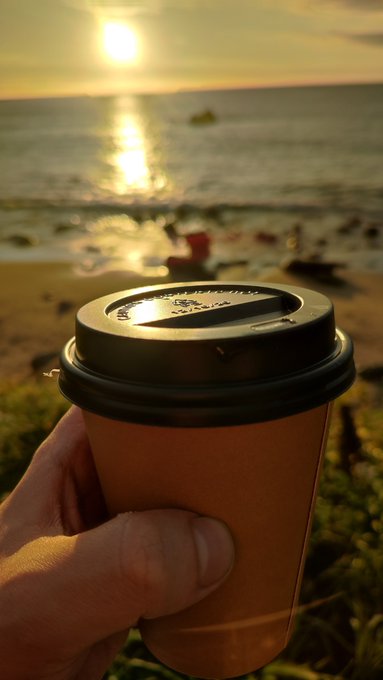 A hand holds a takeaway coffee cup with a black lid against a golden sunset over the ocean, with a small boat visible on the horizon. The peaceful beach scene complements the post's theme of daily discipline and showing up consistently, even when there's no immediate reward to show for it.