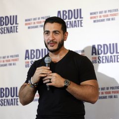 A man with a beard and dark t-shirt speaks into a microphone in front of a backdrop reading 'ABDUL for SENATE - MONEY AND/OR PROTEST MONEY TO YOUR PERMIT MICHIGAN FINE ME.'
