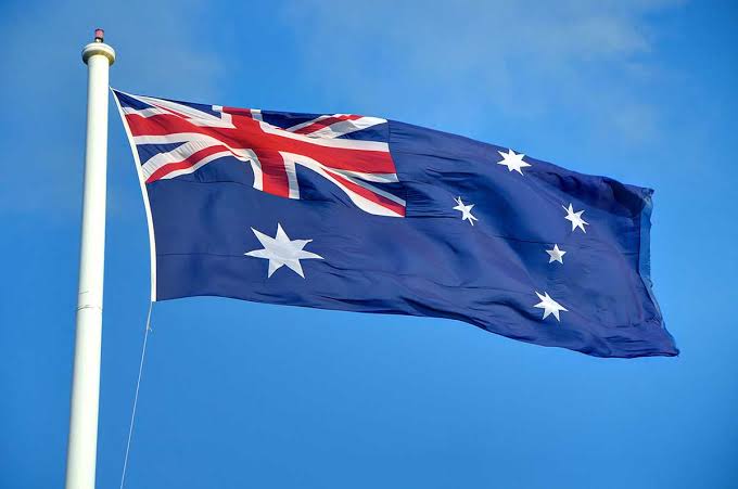 Australian flag waving on a white flagpole against a clear blue sky, featuring the Union Jack, the Commonwealth Star, and the Southern Cross constellation.