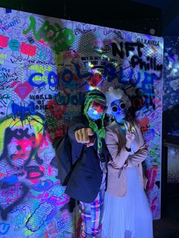 Two people pose in front of a colorful graffiti wall featuring NFT artwork, wearing sunglasses and face masks under UV/blacklight illumination.